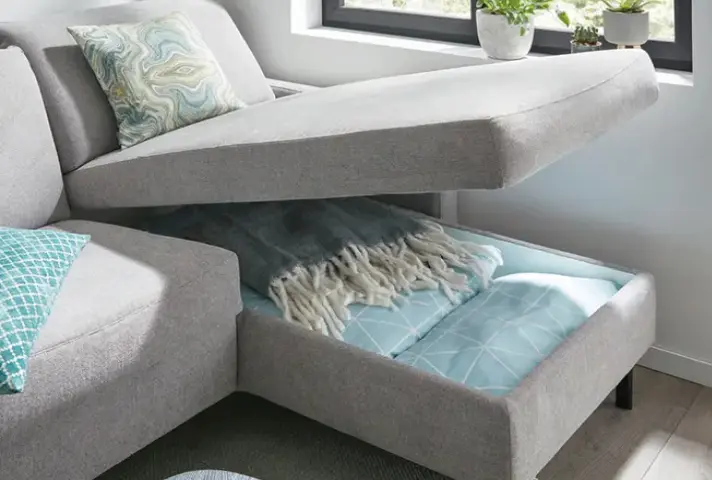 Graues Ecksofa mit hochgeklappter Sitzfläche, im Stauraum darunter Decke und Bettdecke in Türkis, daneben Fensterbank mit Pflanzen und Teppichboden.